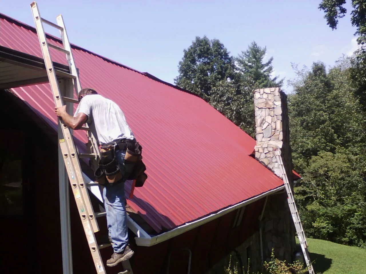 Roofer on a ladder beside a red metal roof and stone chimney — Soffit & Fascia Repair by Roofing Brothers in Fort Wayne